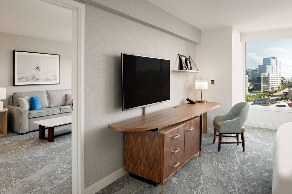 Un salon moderne avec un téléviseur à écran plat, un canapé gris agrémenté de coussins bleus, une console en bois, un fauteuil gris et une vue sur la ville par une grande fenêtre.