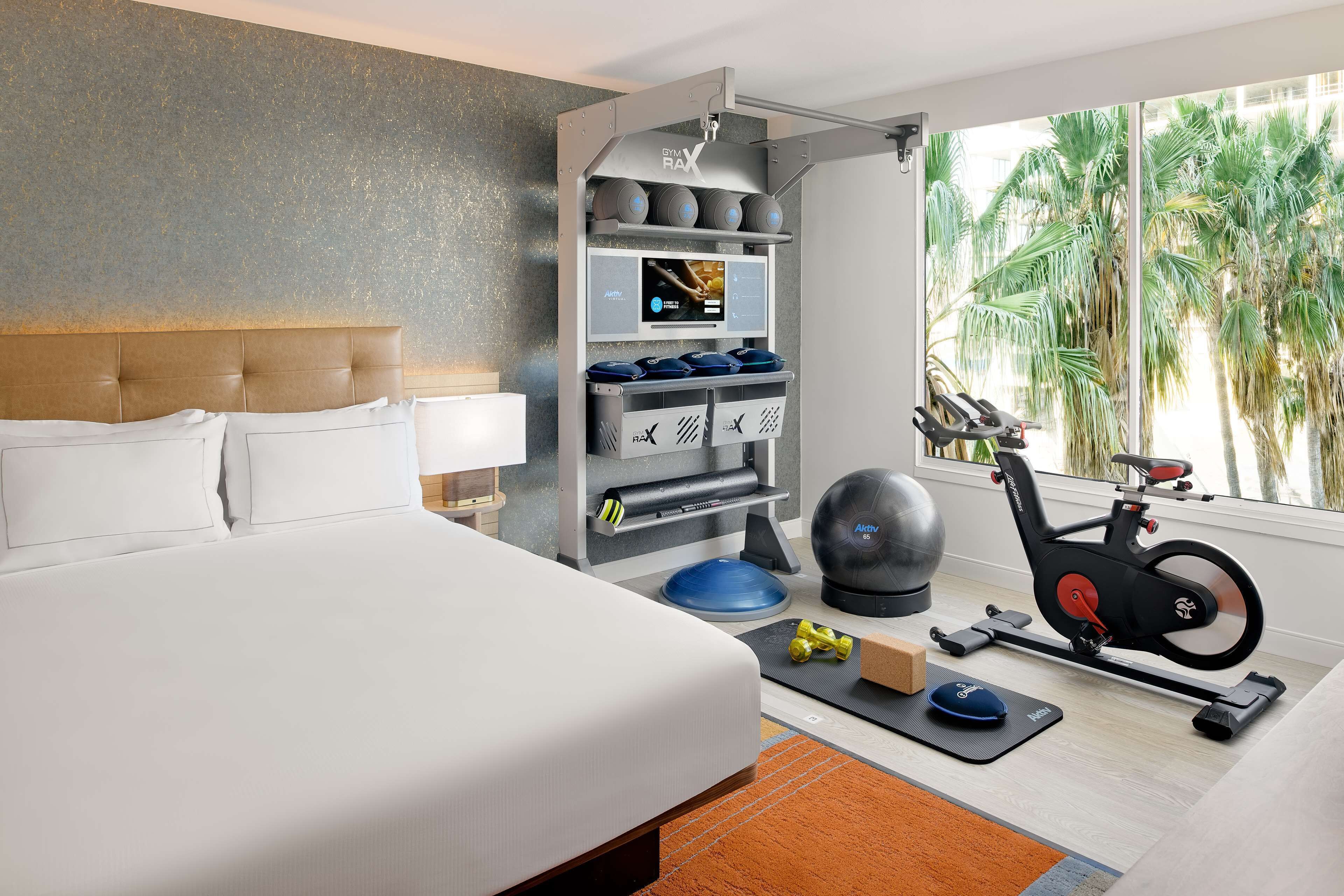 Habitación de hotel con cama, equipo de ejercicio, pantalla digital y vistas a palmeras.
