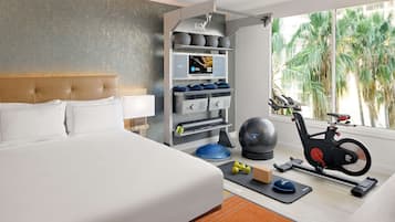 Habitación de hotel con cama, equipo de ejercicio, pantalla digital y vistas a palmeras.