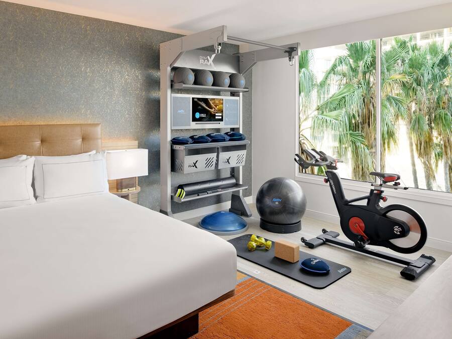Habitación de hotel con cama, equipo de ejercicio, pantalla digital y vistas a palmeras.