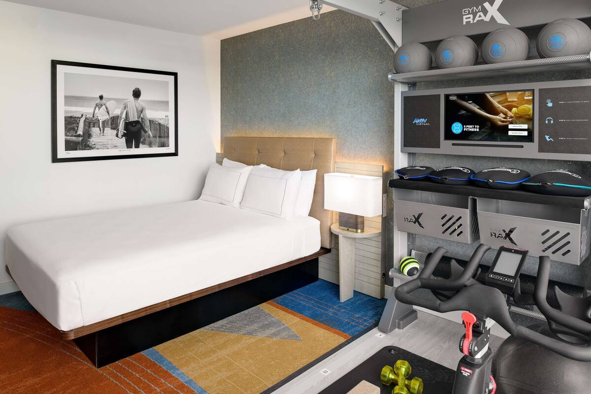 Une chambre d’hôtel avec un lit, un espace dédié à l’exercice physique comprenant des ballons d’exercice et un écran, et un tableau encadré accroché au mur.
