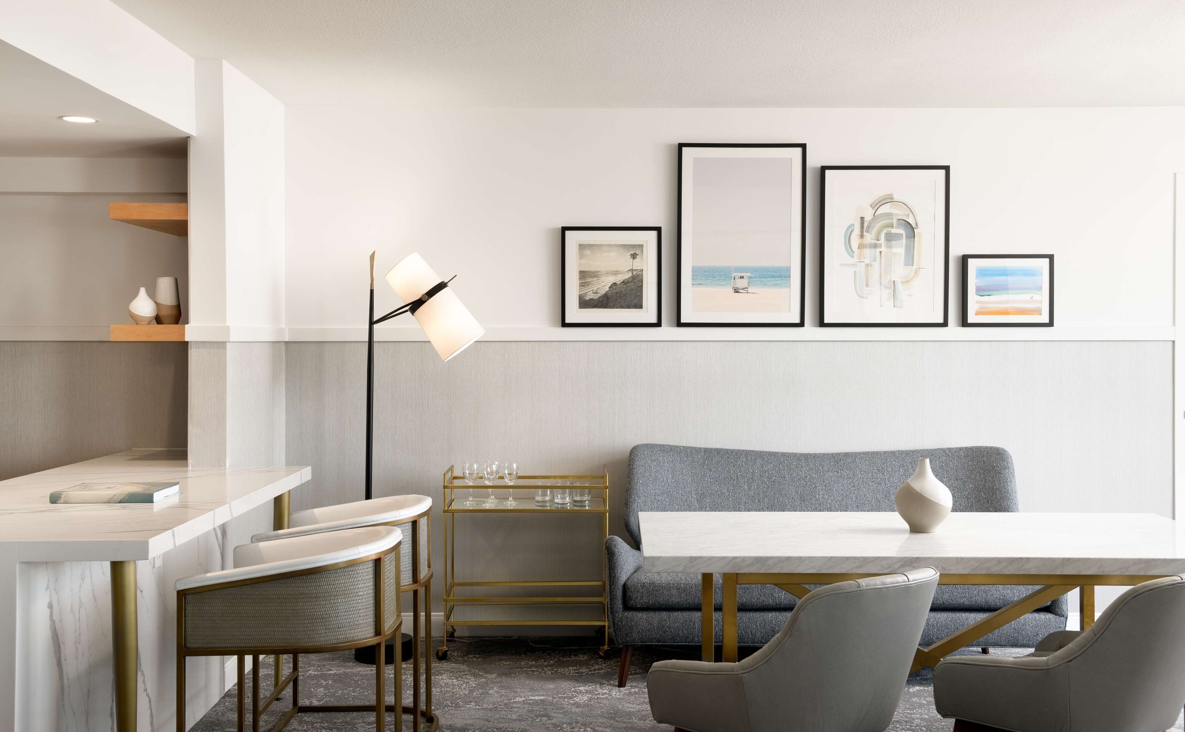A modern dining area with a marble countertop, a high table, and a bench. There are framed artworks on the wall and a floor lamp.