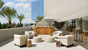 Een terras op het dak met witte banken, poufs en houten bijzettafeltjes, een houten bar en uitzicht op palmbomen en een modern gebouw.