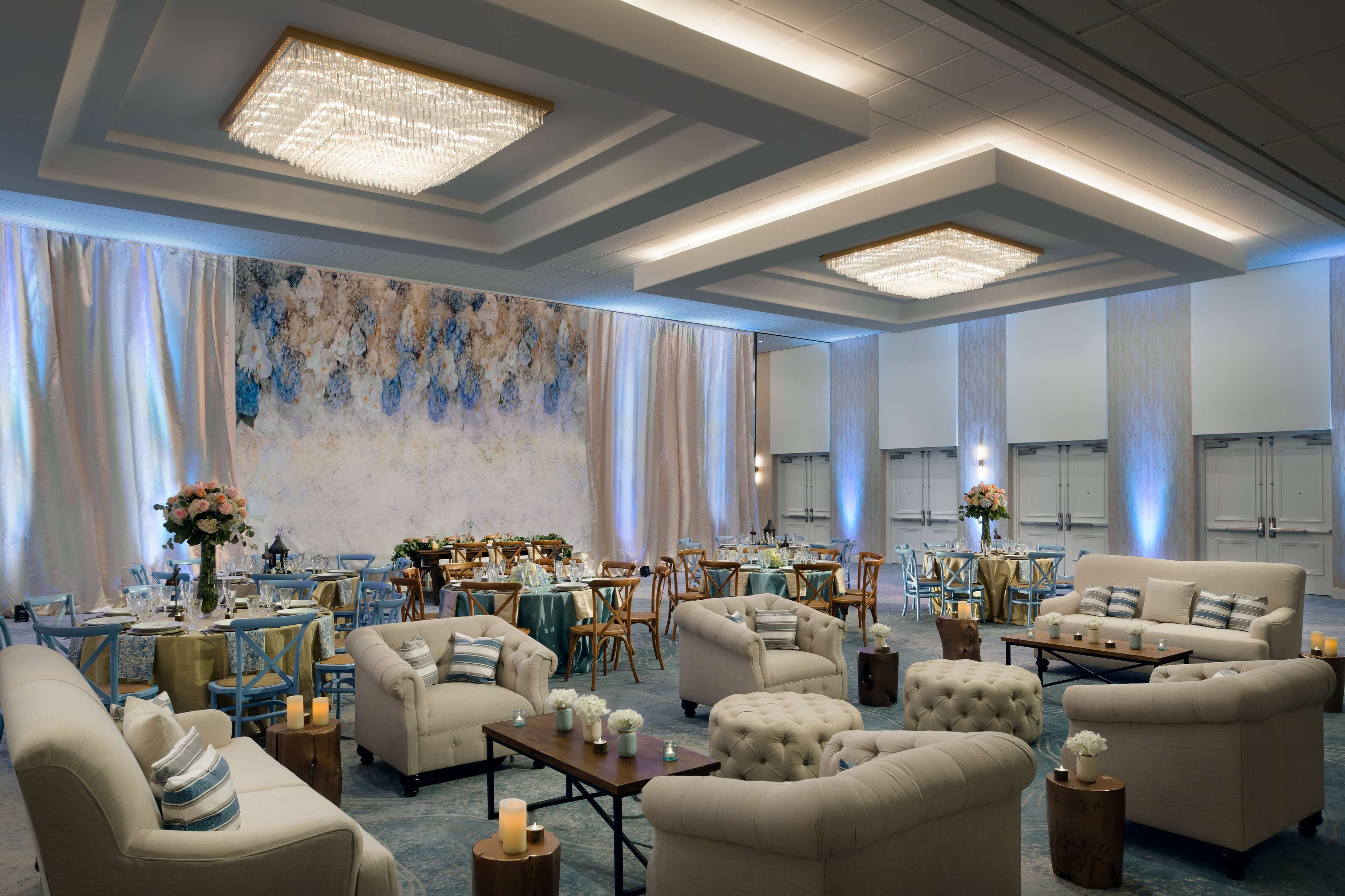 A spacious hotel lobby with a high ceiling, chandeliers, and a large abstract painting on the wall. There are multiple seating areas with sofas and tables, and a dining area in the background.