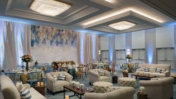 A spacious hotel lobby with a high ceiling, chandeliers, and a large abstract painting on the wall. There are multiple seating areas with sofas and tables, and a dining area in the background.