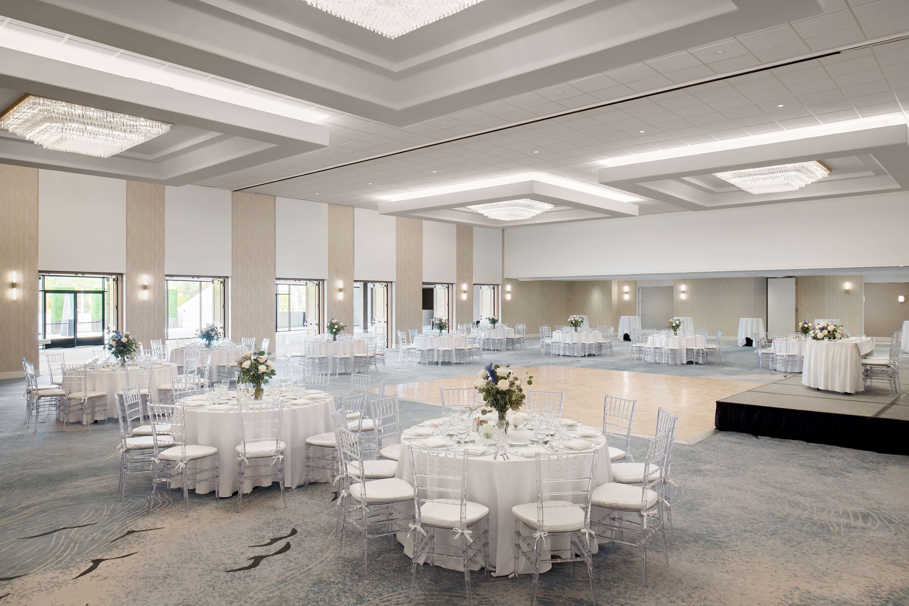 Un amplio salón de banquetes, bien iluminado, con varias mesas redondas dispuestas para eventos, cubiertas con manteles blancos y sillas transparentes.