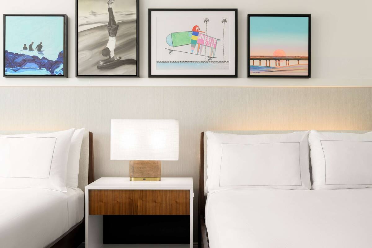 Une chambre d’hôtel avec deux lits, une table de chevet avec une lampe et trois œuvres d’art encadrées accrochées au mur.