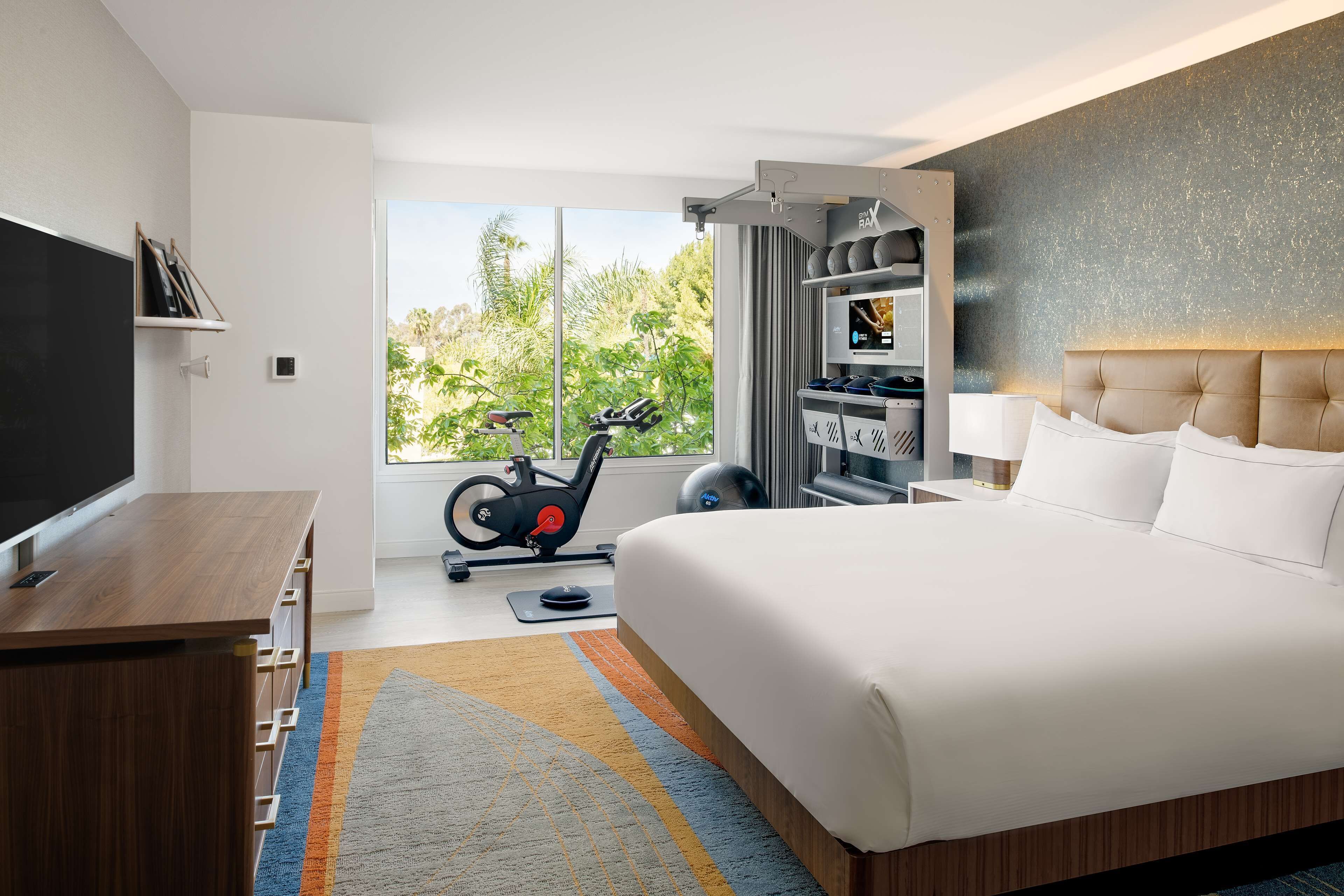 A modern hotel room with a large bed, a desk, a television, and a view of greenery outside.