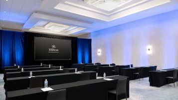 A conference room with a large screen displaying the Hilton logo, multiple rows of tables with chairs, water bottles on each table, and a chandelier on the ceiling.