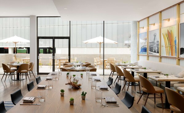 Un restaurant moderne avec des tables et des chaises en bois, de grandes fenêtres et des plantes d’intérieur.
