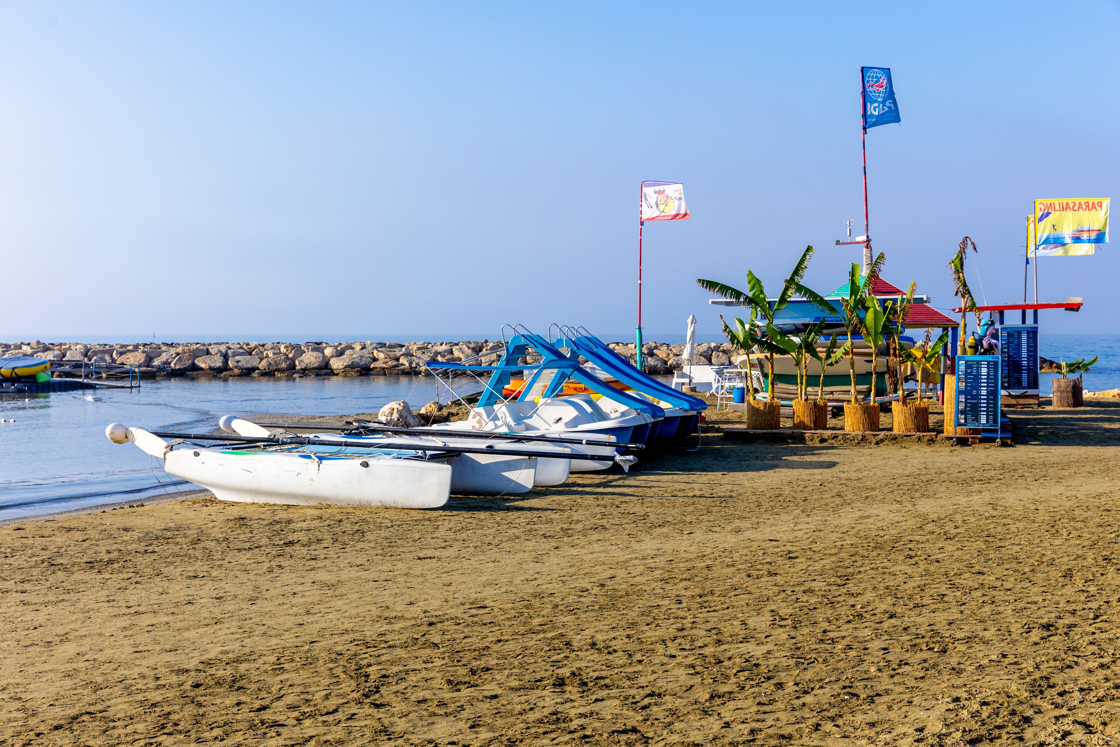 A beach with several boats docked near the shore, a small structure with a red roof, and flags.