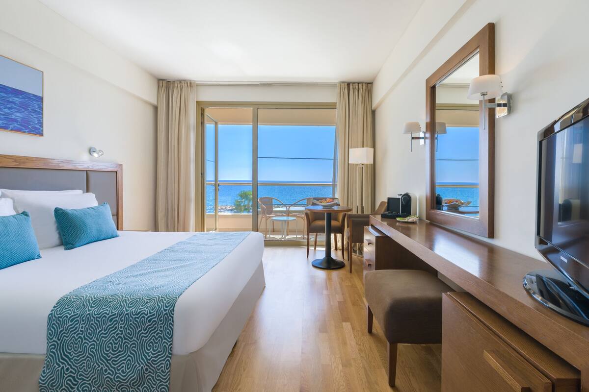 Quarto de hotel com cama grande, escrivaninha, televisão e varanda vista para o mar.