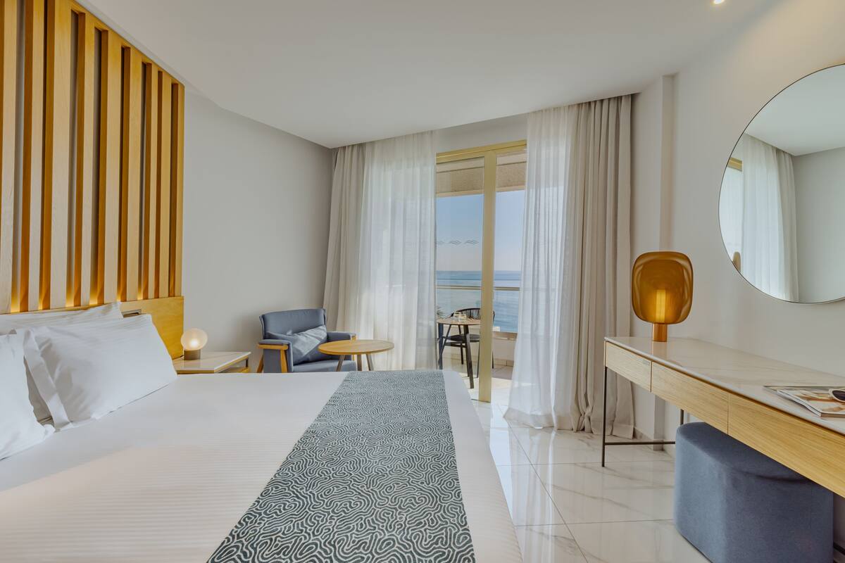 Quarto de hotel moderno com uma cama grande, uma escrivaninha, uma cadeira e vista para o mar.