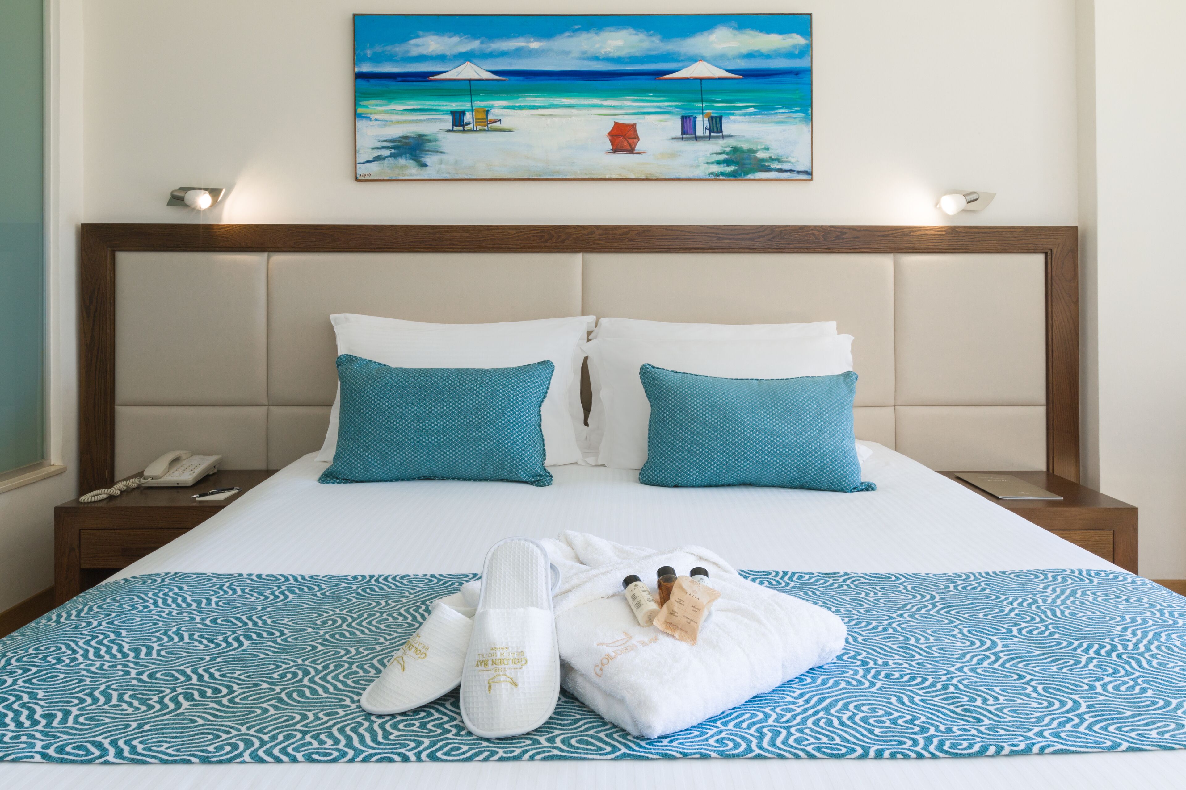 A neatly made bed with a patterned bedspread, two pillows, a pair of slippers, and a towel on a wooden nightstand with a phone.