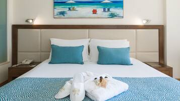 A neatly made bed with a patterned bedspread, two pillows, a pair of slippers, and a towel on a wooden nightstand with a phone.