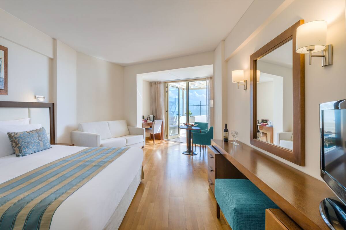 Quarto de hotel com cama, escrivaninha, cadeira, televisão e uma janela grande.