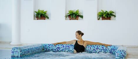 A woman in a black swimsuit in a pool with blue mosaic tiles, surrounded by white columns and potted plants.
