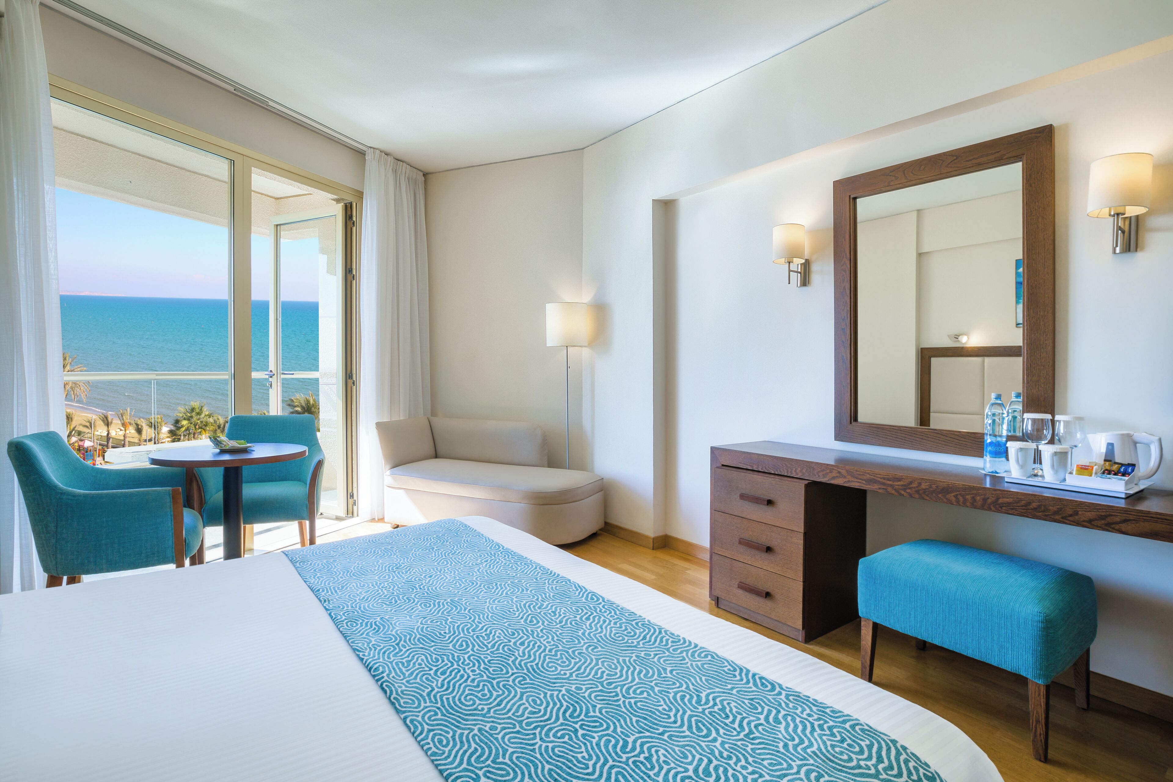 Habitación de hotel con cama, escritorio, silla, banco, un espejo grande y vista al océano.