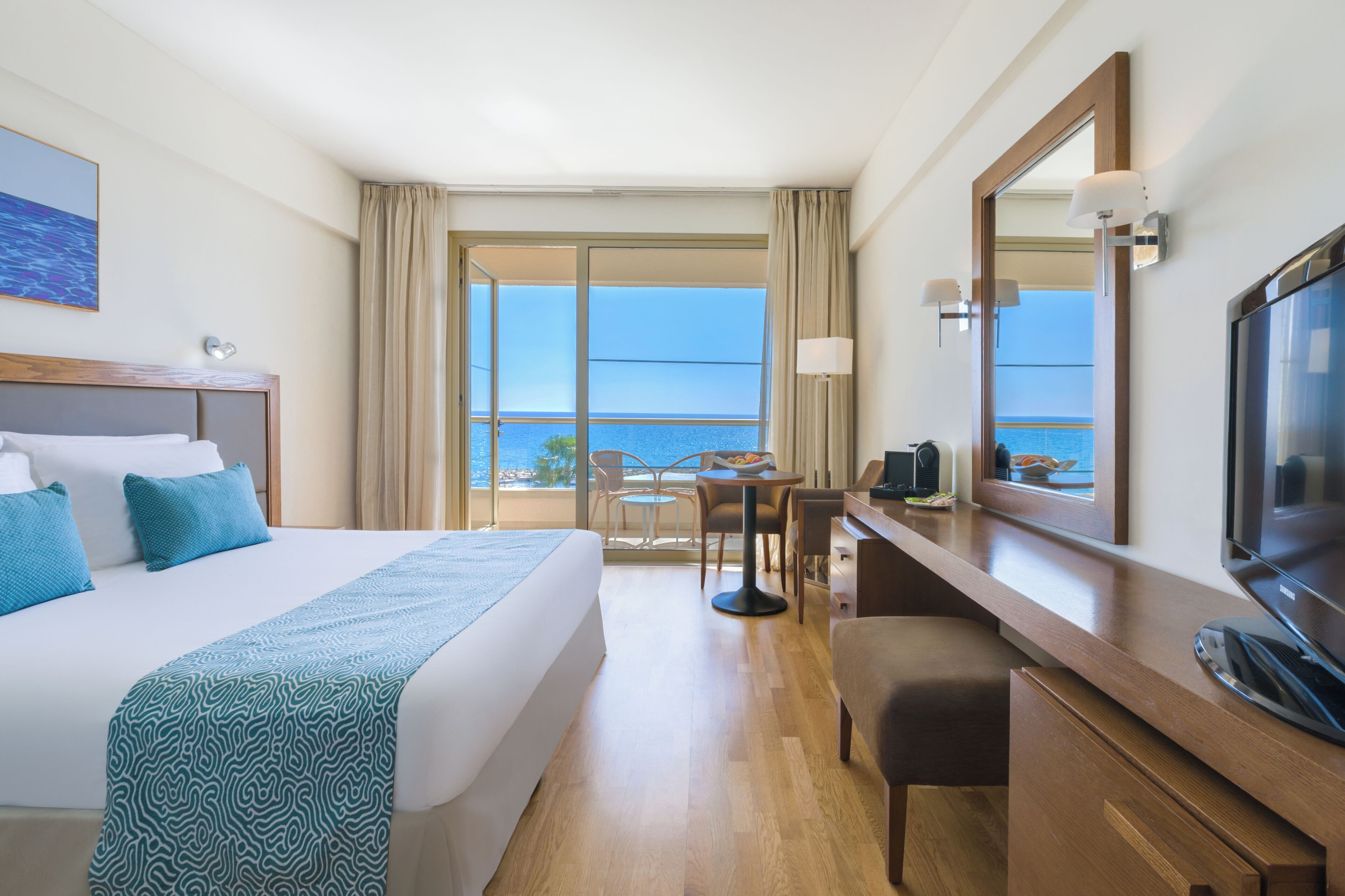 Habitación de hotel con una cama grande, un escritorio, televisión y un balcón con vista al mar.