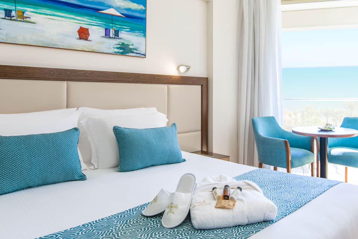 Quarto de hotel com uma cama grande, almofadas azuis e brancas, colcha estampada, pintura de uma cena de praia e uma varanda com cadeiras e uma mesa.