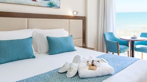 A hotel room with a large bed, blue and white pillows, a patterned bedspread, a painting of a beach scene, and a balcony with chairs and a table.