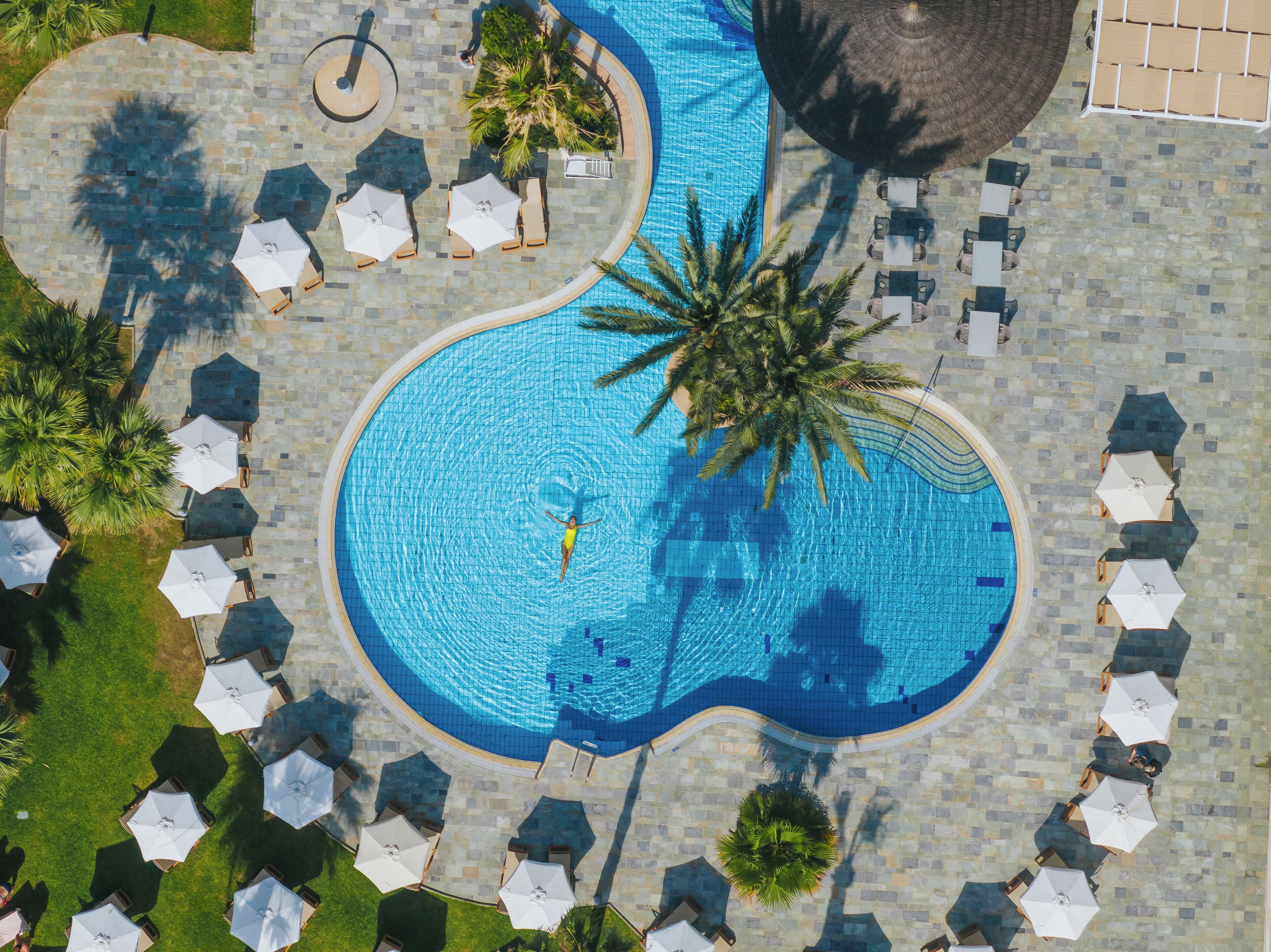 Eine Luftaufnahme eines Swimmingpools mit einem gitarrenförmigen Design, umgeben von Liegestühlen und Sonnenschirmen.
