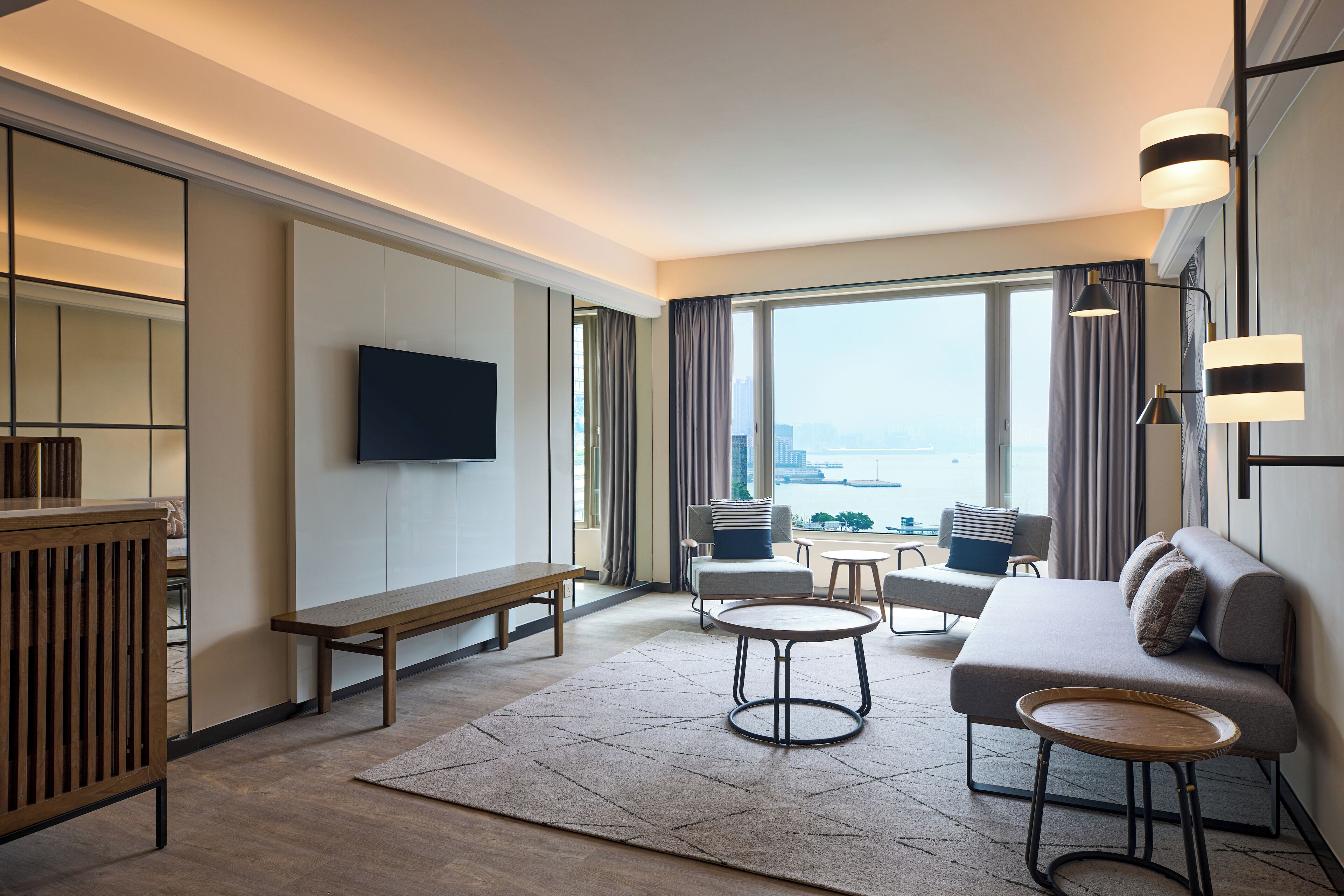 A modern hotel room with a large window, a flat-screen TV, a sofa, a coffee table, and a bench.