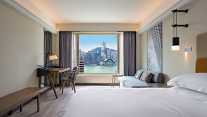 A hotel room with a large bed, a desk, a chair, a small table, and a view of a city skyline.