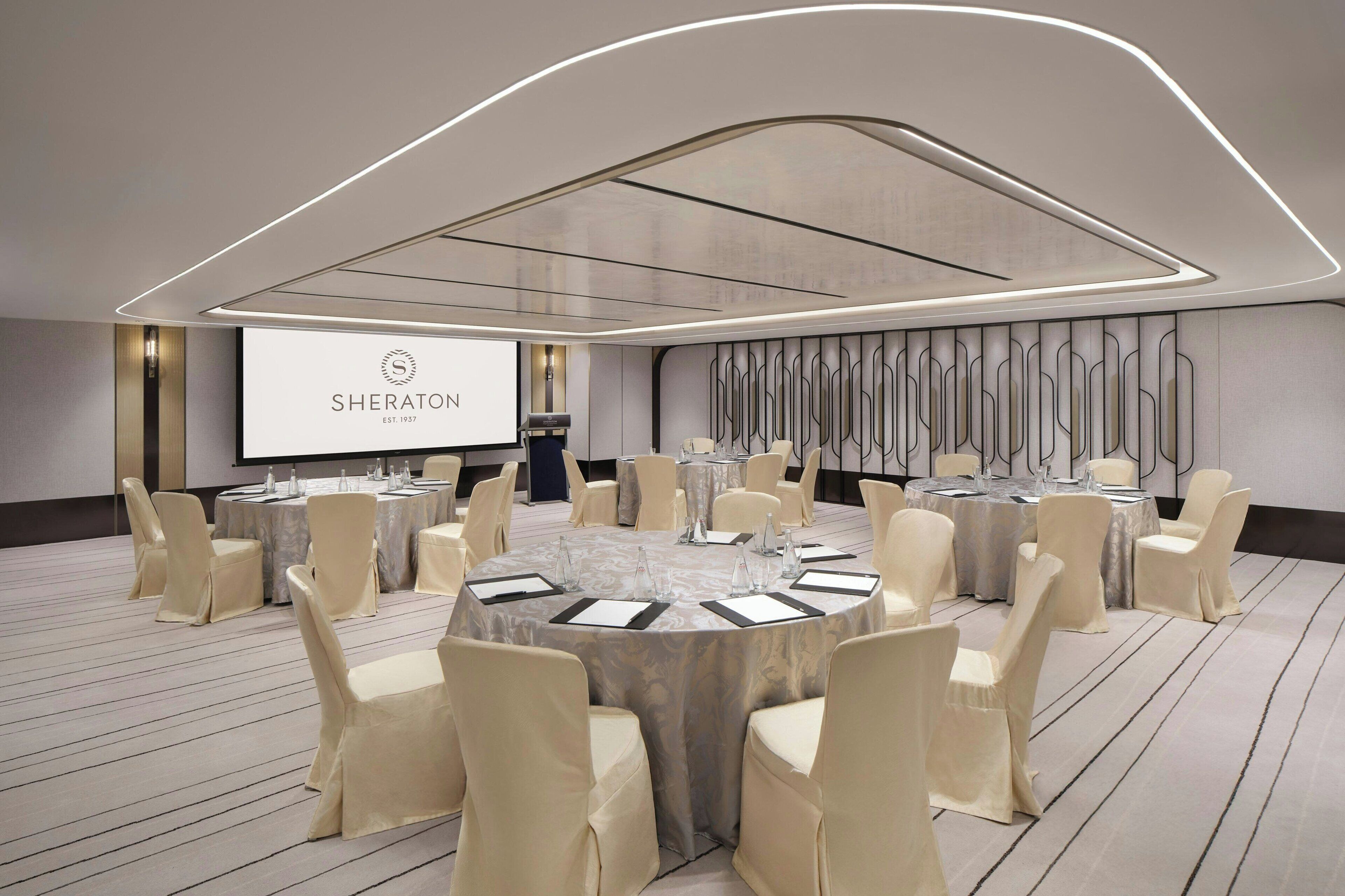 A conference room with round tables, beige chairs, and a large screen displaying 'SHERATON'.