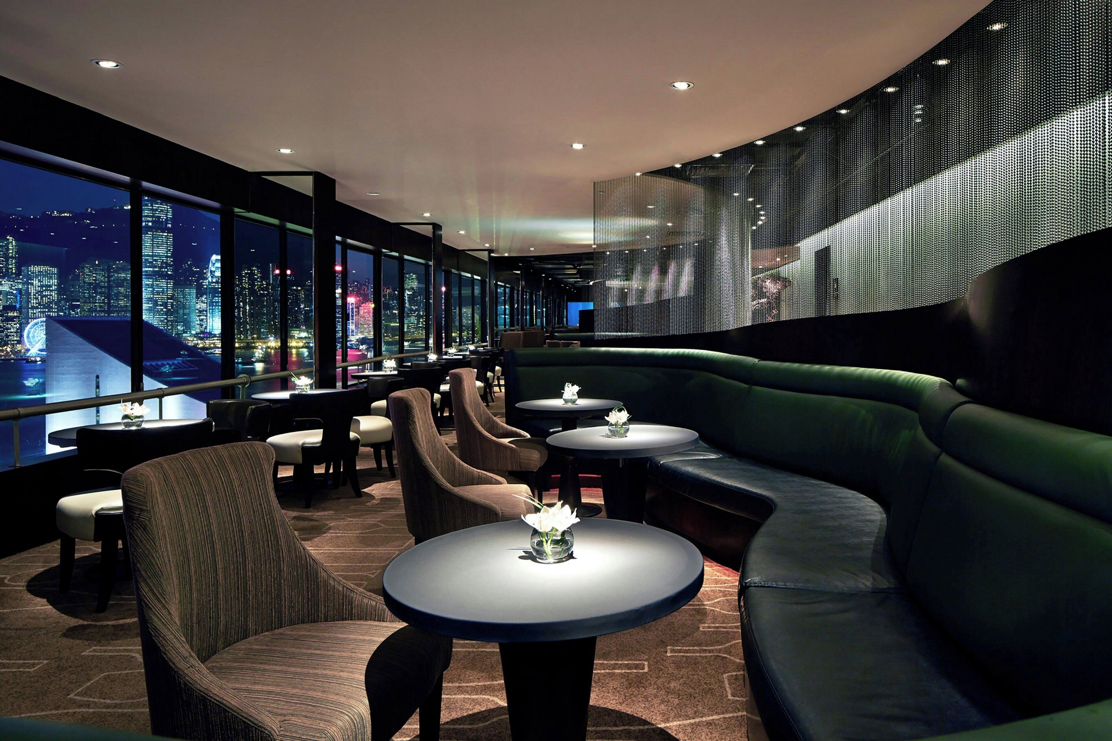 A modern lounge area with seating and tables, large windows offering a city view, and a decorative wall.