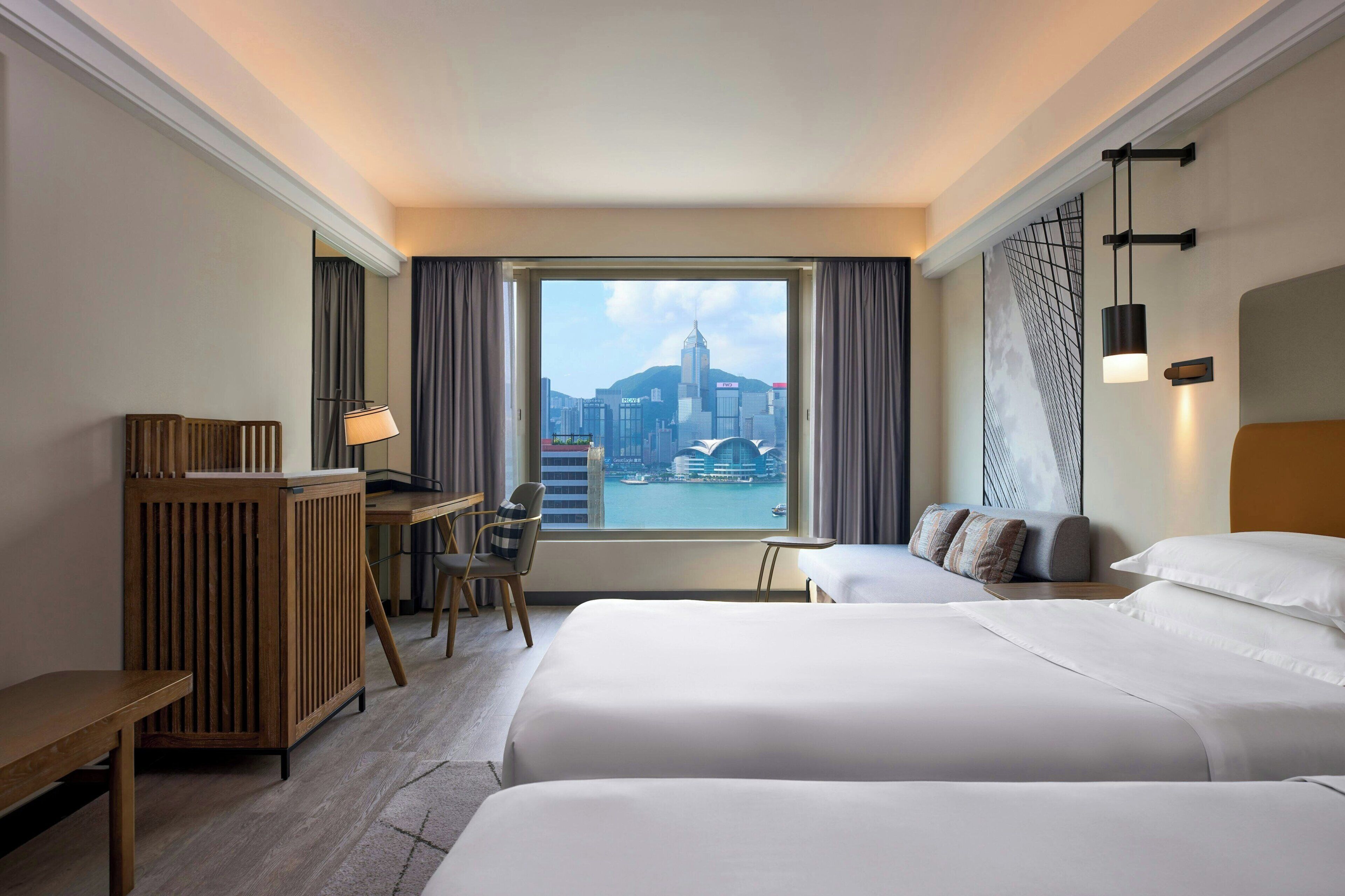 Quarto de hotel com duas camas, uma escrivaninha e uma cadeira. Tem uma janela grande com vista para a cidade.