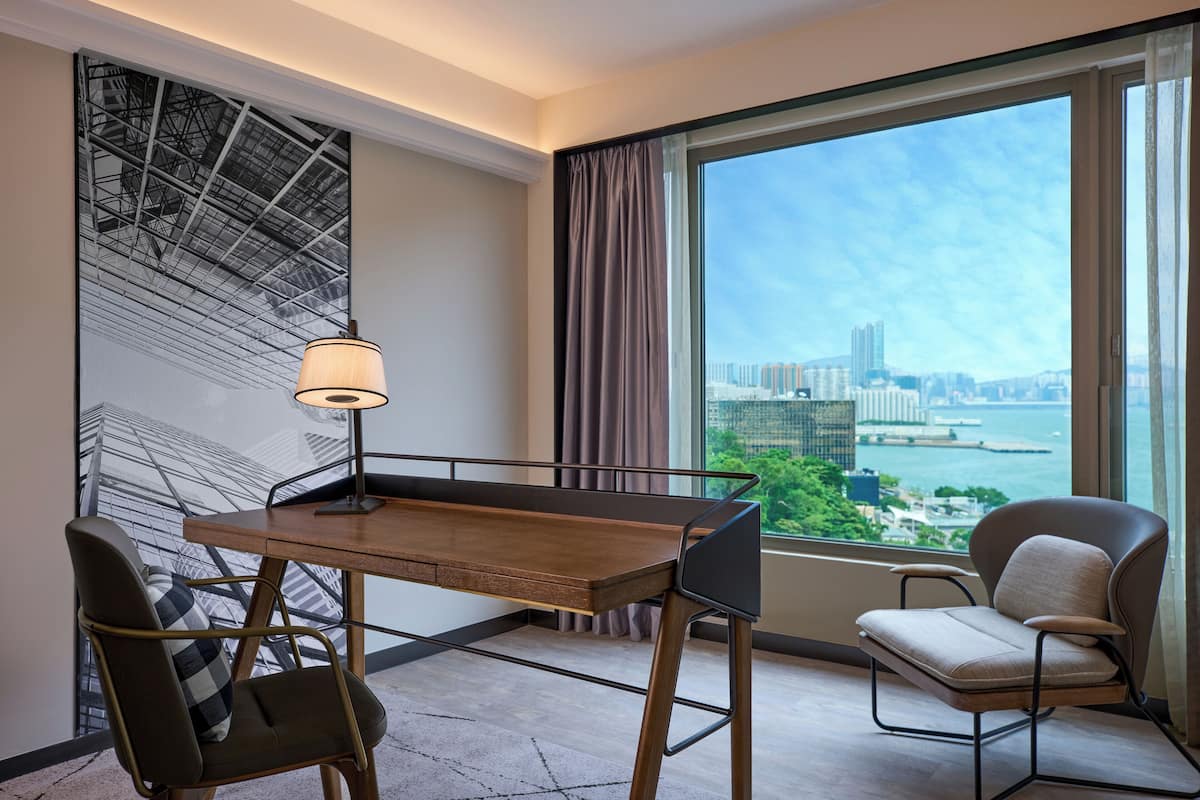 A modern hotel room with a large window offering a city view, a wooden desk with a lamp, and two chairs.
