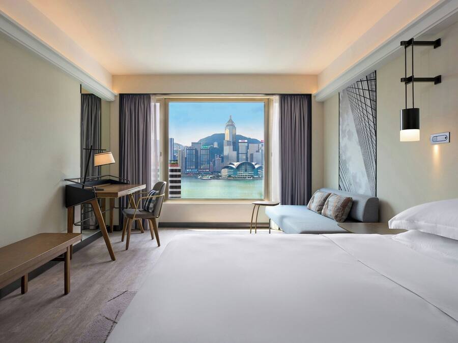 Habitación de hotel con una cama grande, un escritorio, una silla, una mesita y vistas a un paisaje urbano a través de un ventanal amplio.