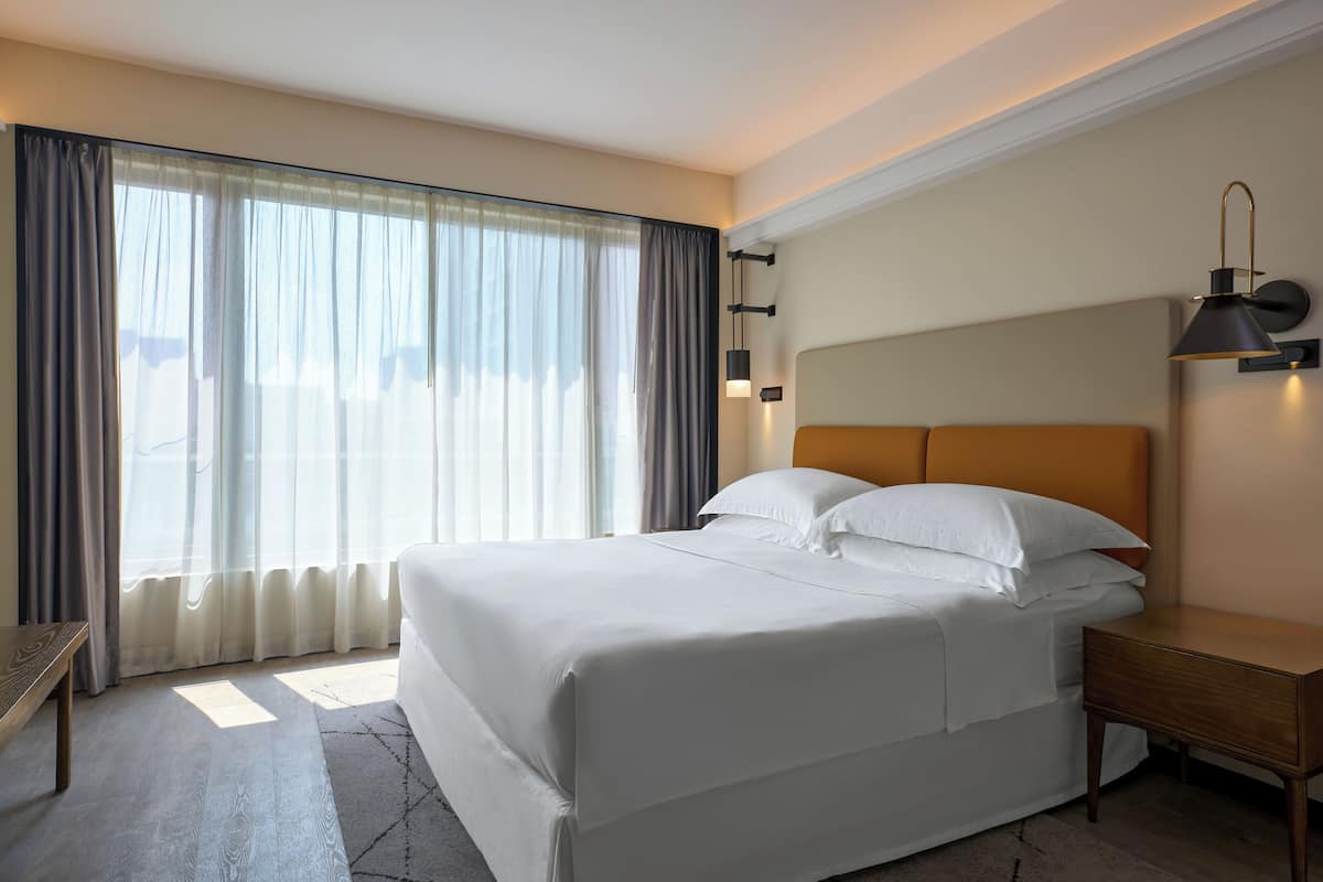 A hotel room with a large bed, a bedside table, a lamp, and a view of the outdoors through sheer curtains.