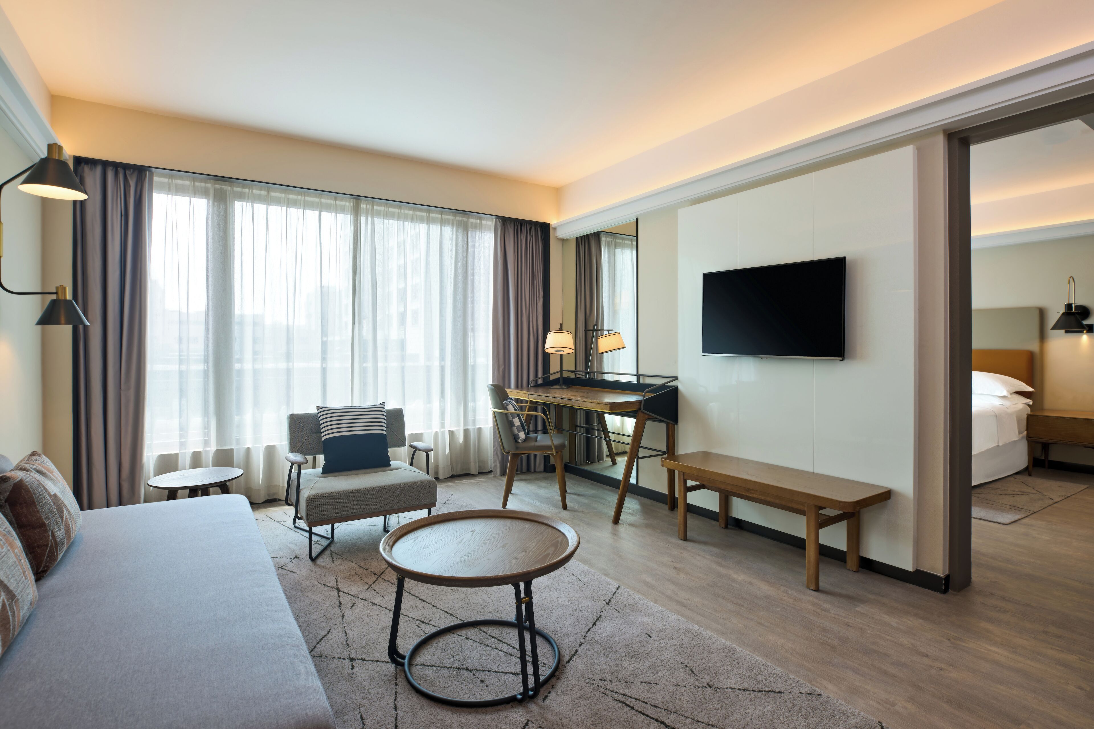 A modern hotel room with a sofa, a round table, a desk, a TV, and a bed.