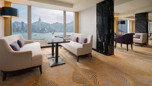 A modern hotel room with a large window offering a city view, a plush sofa, a dining table, and a marble accent wall.