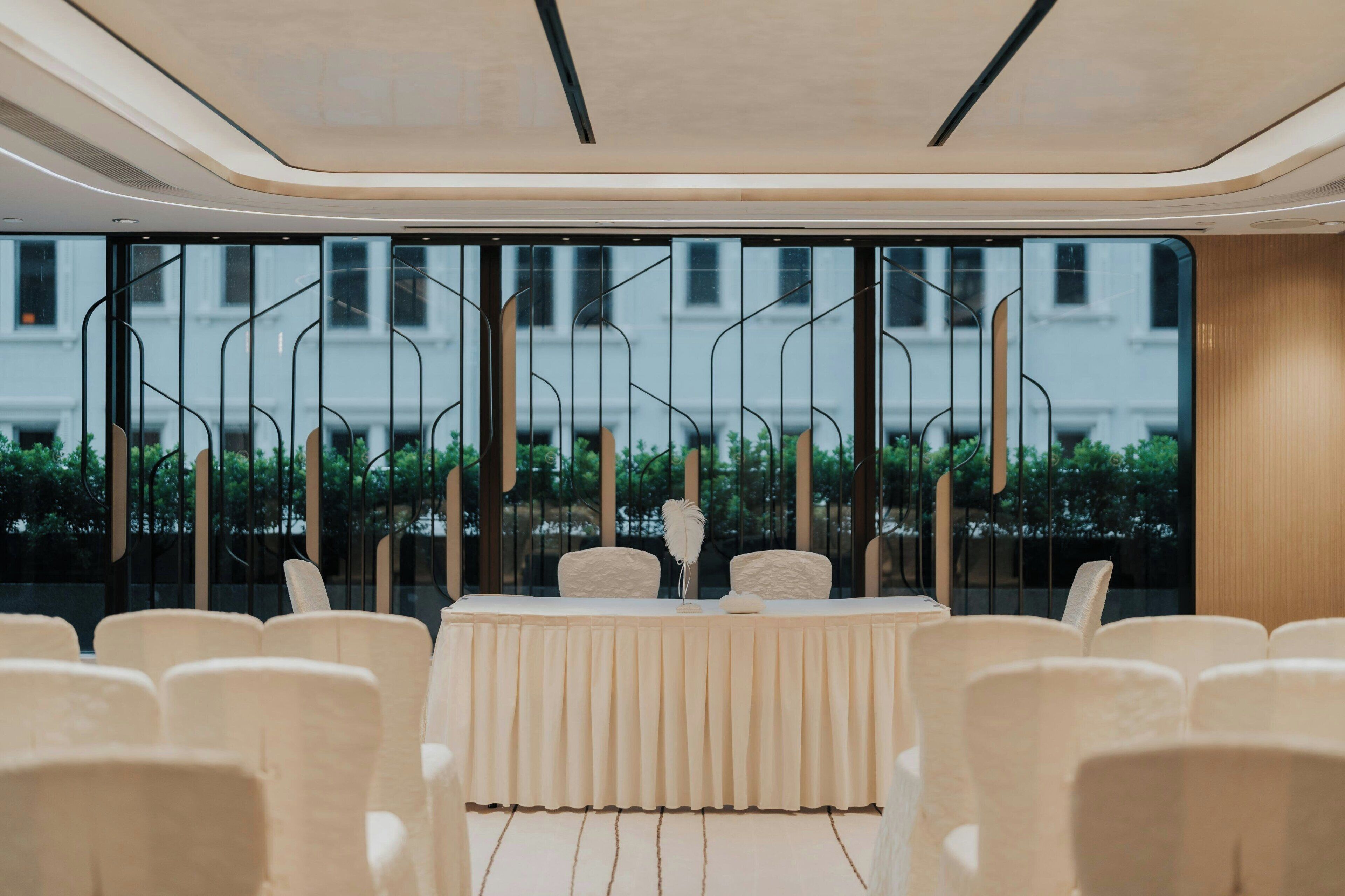 A conference room with a long table, white chairs, and a view of buildings through large windows.