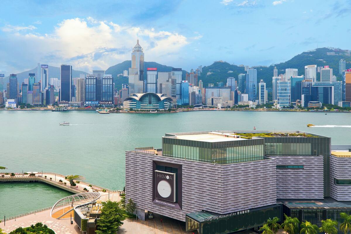 A modern building with a large HKMOA sign, situated near a waterfront with a city skyline in the background.