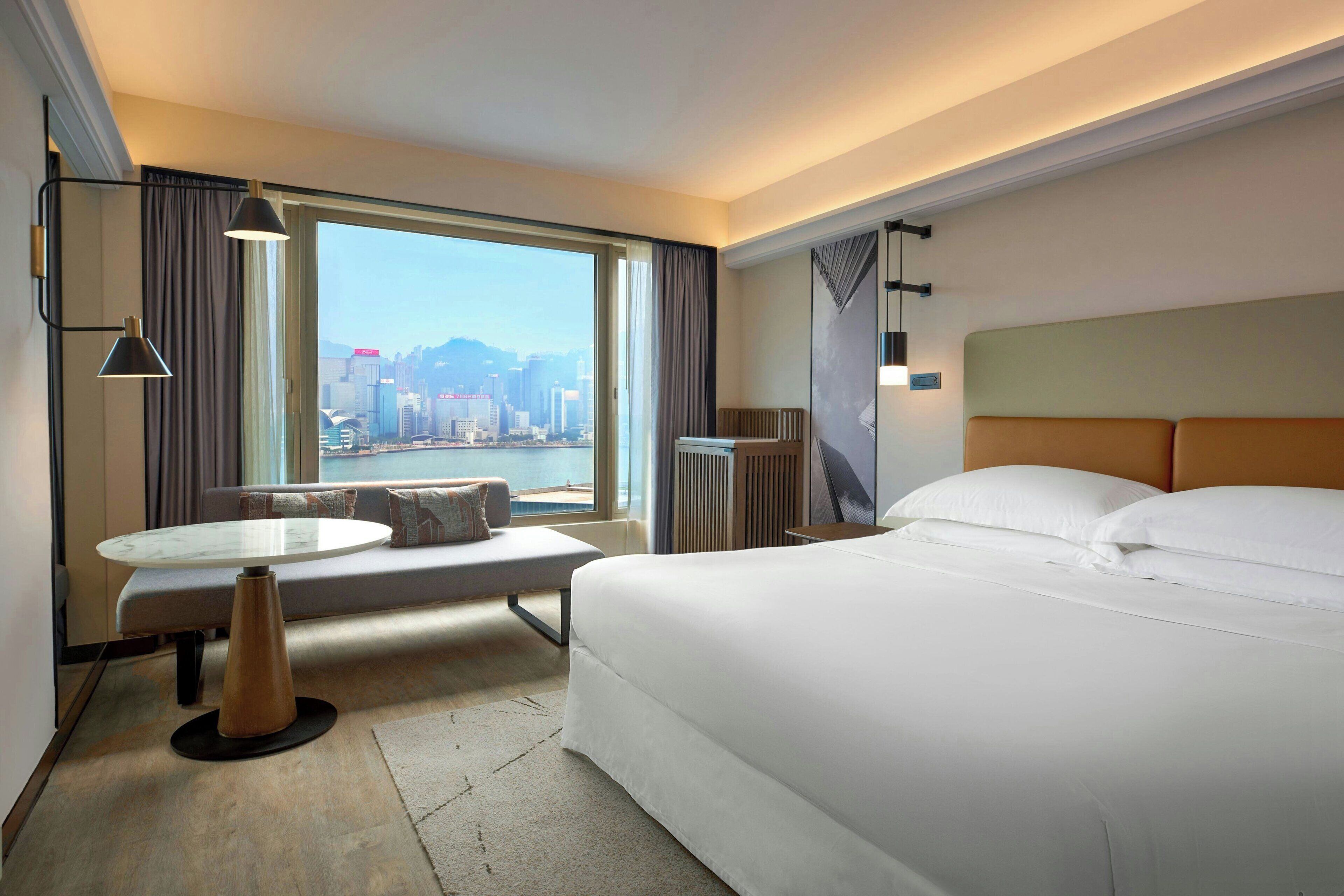 A hotel room with a large bed, a round table, a bench, and a view of a cityscape through a large window.
