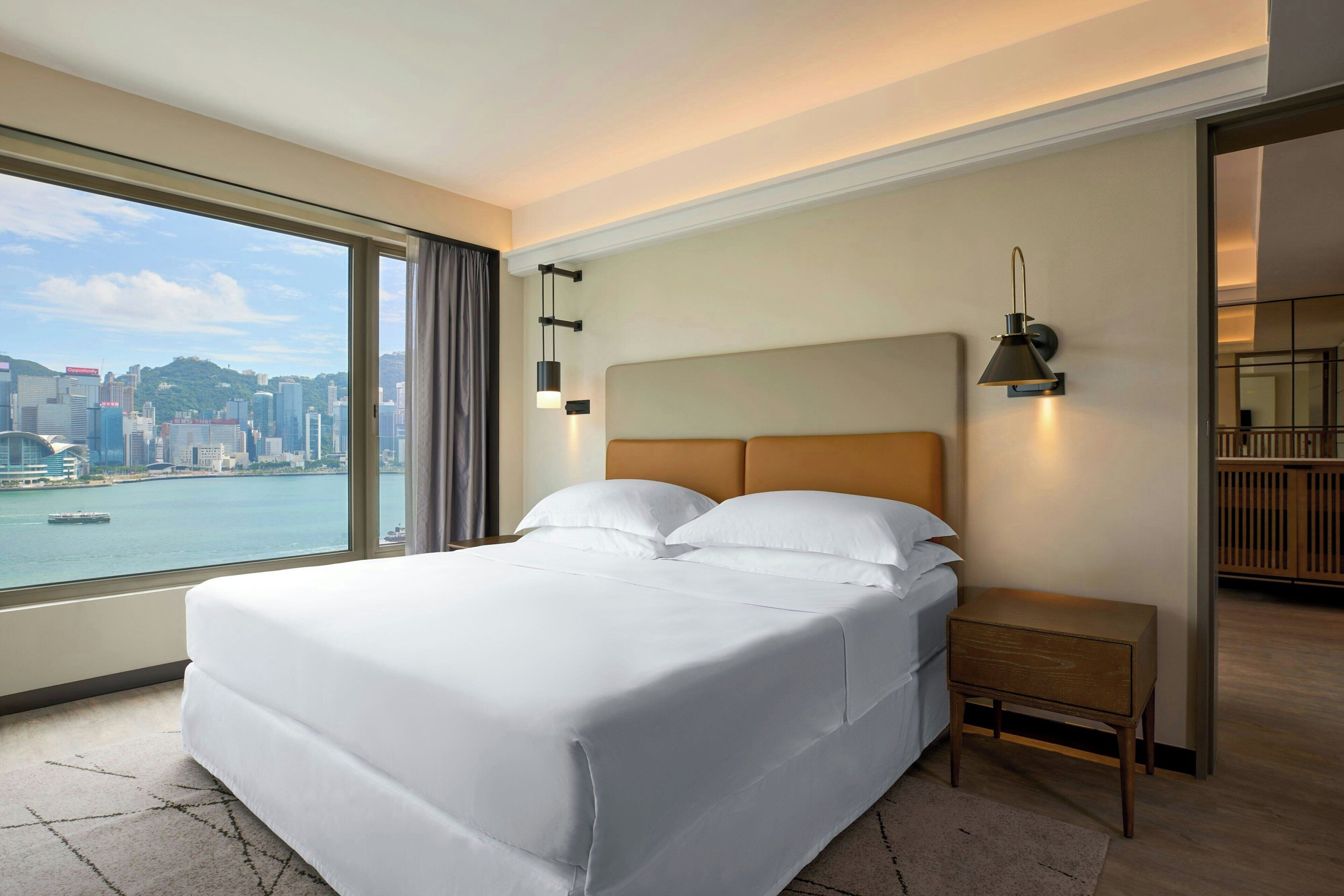 A hotel room with a large bed, a nightstand, a lamp, and a view of a cityscape and water.