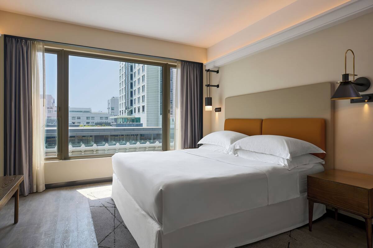 A hotel room with a large bed, a nightstand, a lamp, and a view of the city through large windows.