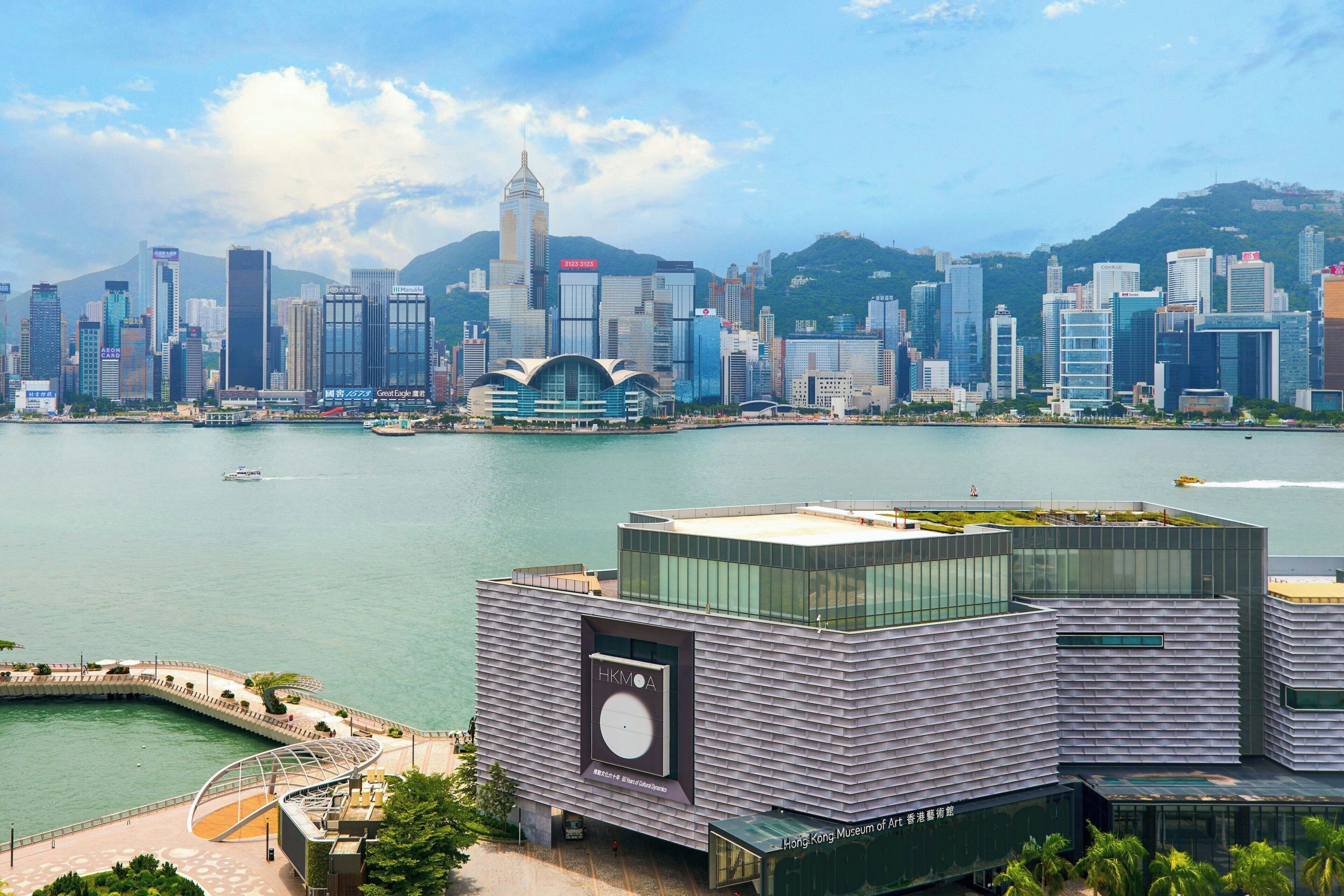 A modern building with a large HKMOA sign, located near a waterfront with a city skyline in the background.