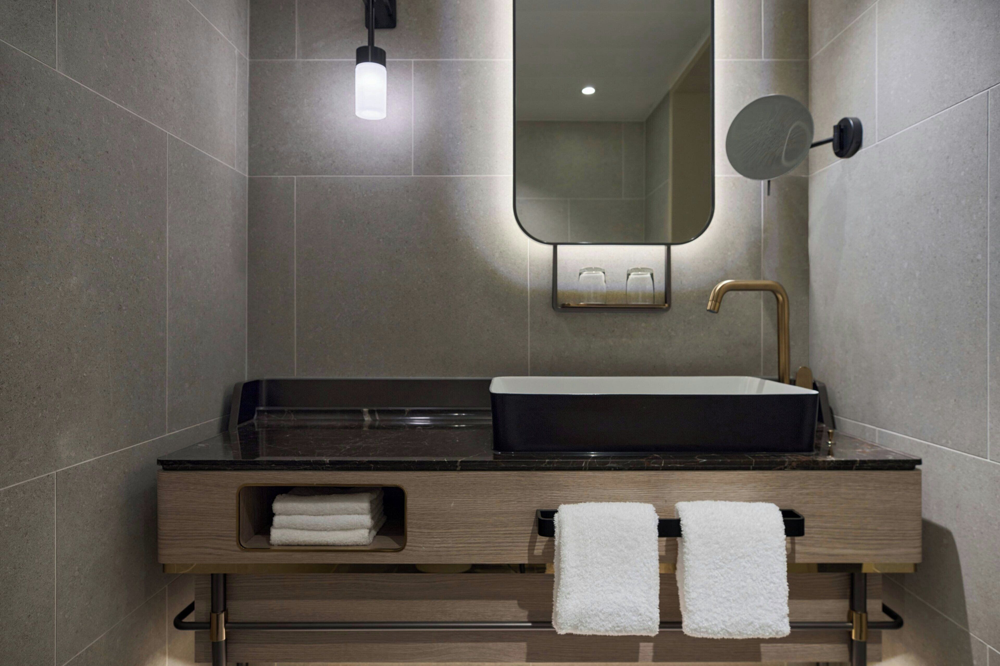 A modern bathroom with a black sink, gold faucet, and a mirror above it. There are two towels hanging on hooks and a small shelf with additional towels.