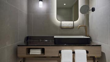 A modern bathroom with a black sink, gold faucet, and a mirror above it. There are two towels hanging on hooks and a small shelf with additional towels.