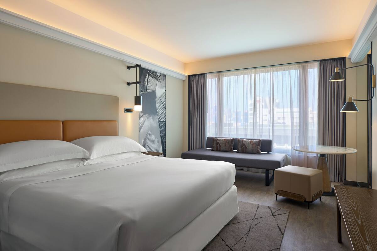 A modern hotel room with a large bed, a seating area with a sofa and ottoman, a small table, and a large window with curtains.