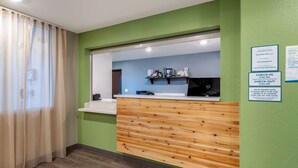 A reception area with a wooden counter, a window with sheer curtains, and a green wall with posted check-in and check-out times.