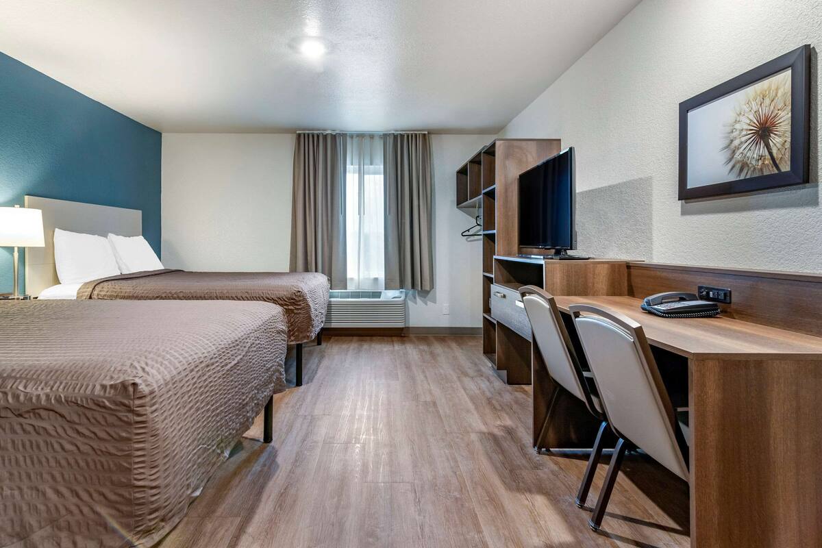 A hotel room with two beds, a desk, a chair, a TV, and a large window with curtains.