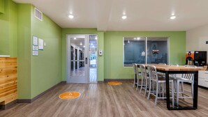 A modern indoor space with a green wall, a wooden bench, a reception desk, and a dining area with chairs and a table.