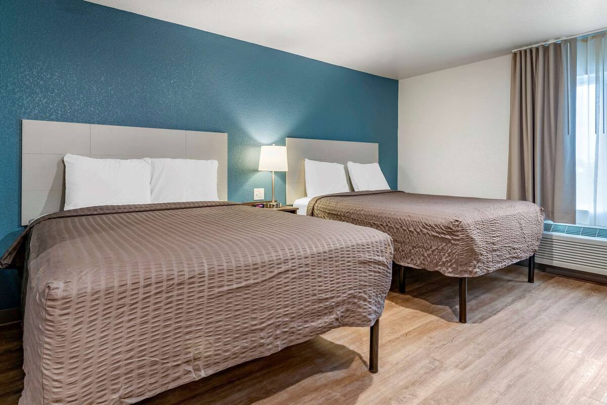 A hotel room with two beds, a blue accent wall, a window with curtains, and a bedside table with a lamp.