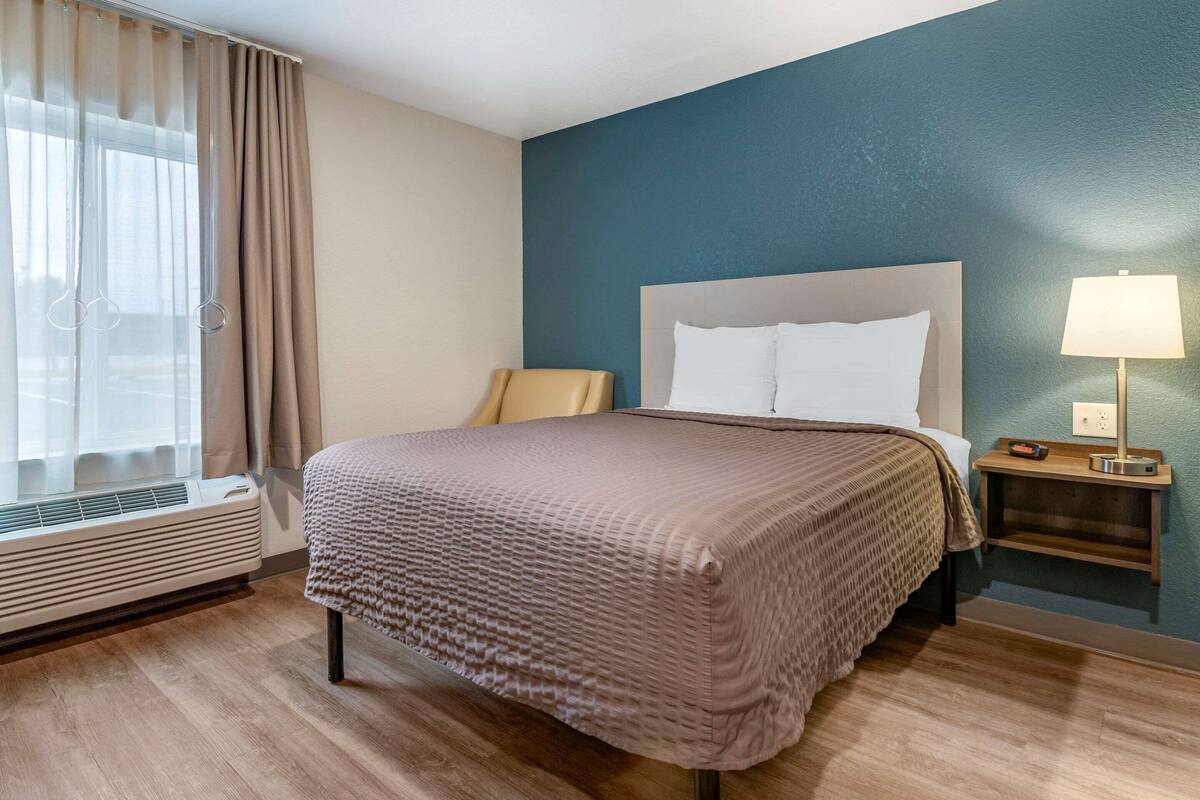A hotel room with a bed, a nightstand, a lamp, a window with curtains, and an air conditioning unit.